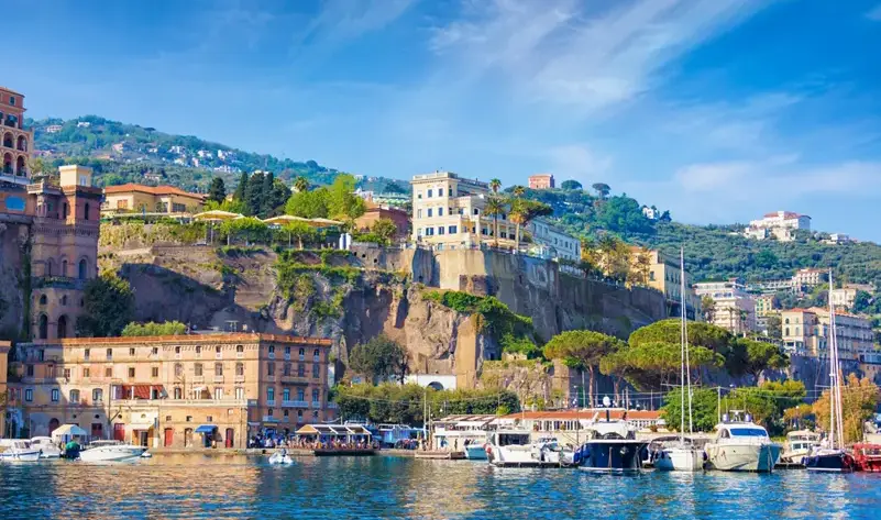 Harbor view of a Mediterranean town with boats docked along a rocky waterfront and hillside homes.