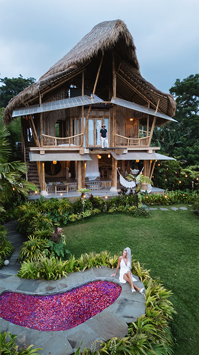 Two-story Thatched Bamboo Villa in a Tropical Garden; a Couple in White Stands on the Upper Balcony While a Woman in White Sits by a Pink-petaled Pool.