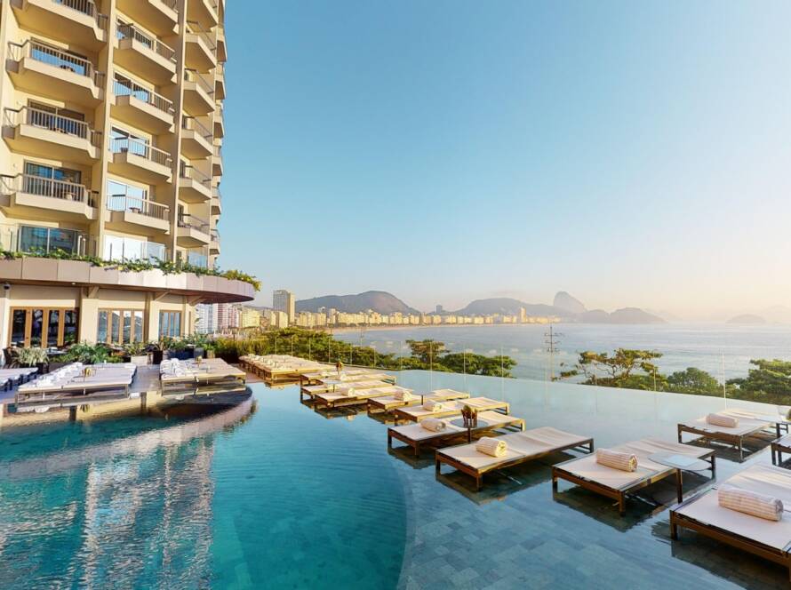 High-rise Hotel with Multiple Balconies Beside an Infinity Pool and a Row of White Loungers Facing the Sea.