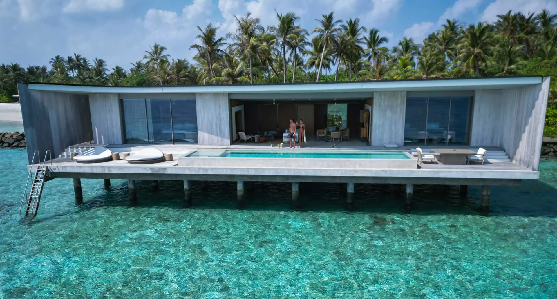 Modern overwater villa on stilts with a central pool deck, glass walls, and palm trees in the background.