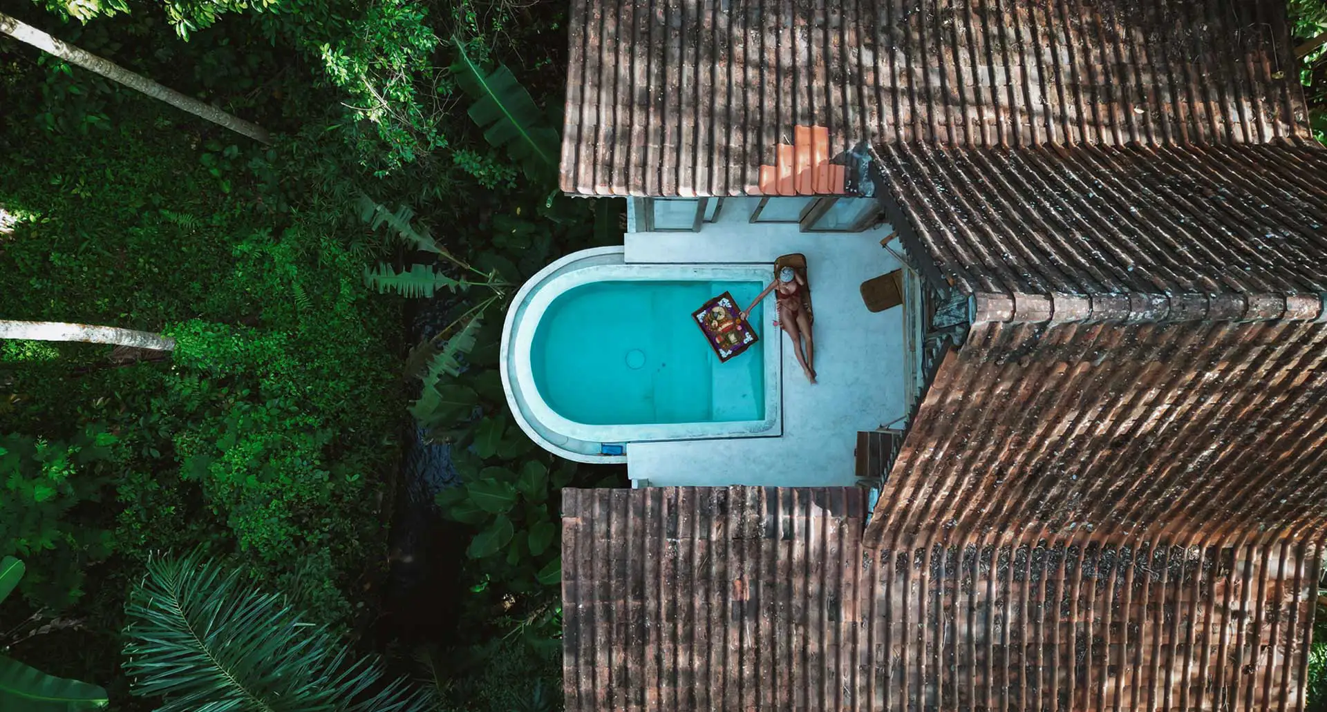 Aerial view of a small turquoise pool beside a weathered roofed structure, surrounded by dense tropical greenery; a person lounges on a chair with a towel.