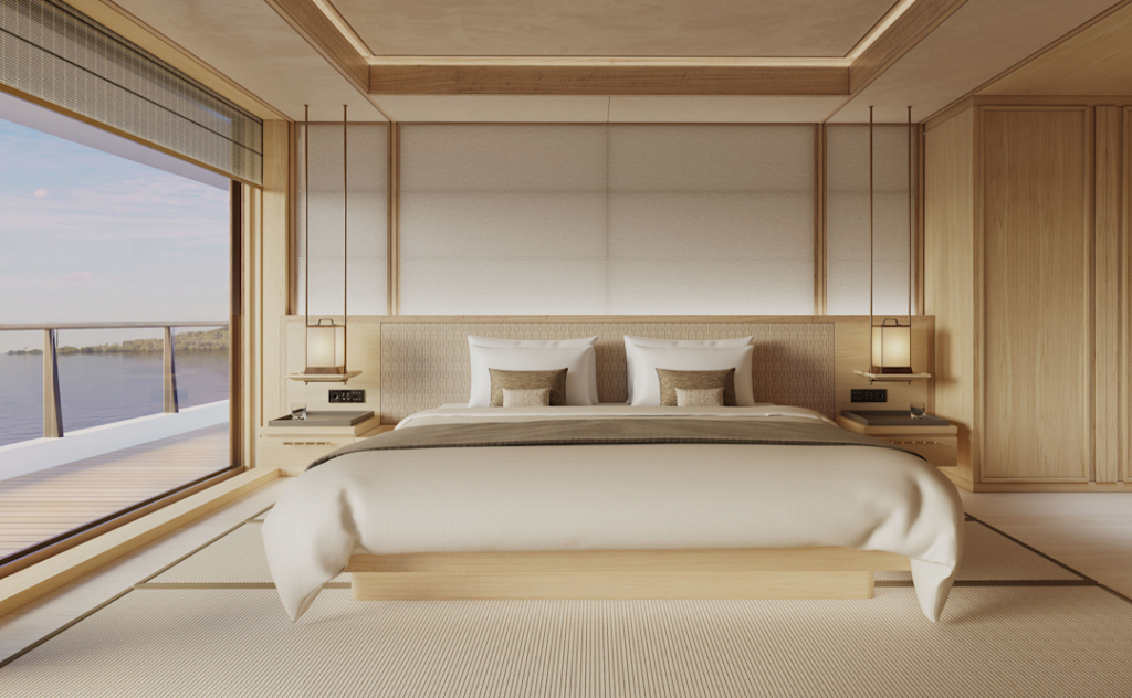 Sunlit hotel bedroom with a low platform bed, white bedding, and twin nightstands, opening to a balcony with a water view on the left.