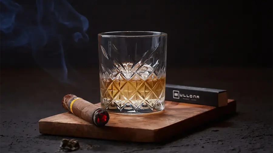 Crystal Whiskey Glass with Amber Liquid on a Wooden Board, Beside a Lit Cigar and a Black Cigarette Pack in the Background