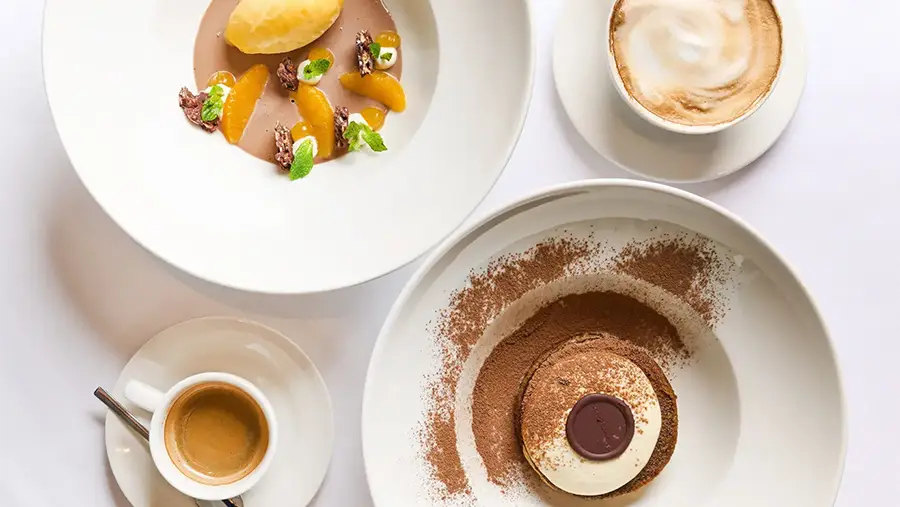 Top-down View of a Chocolate Dessert Ring Dusted with Cocoa, with a Cappuccino and Another Plate Nearby on White Tableware.