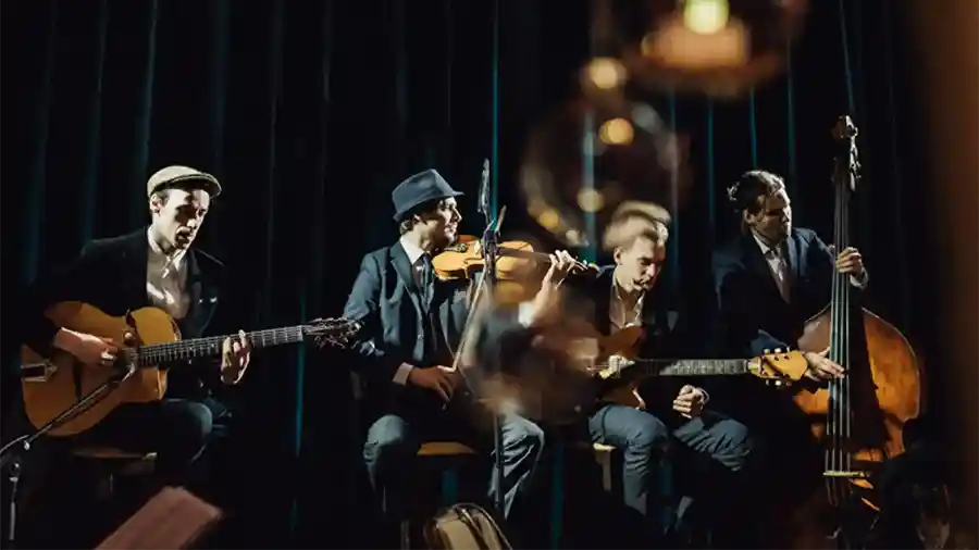 Four Male Musicians in Suits on a Dim Stage; Left Guitarist, Violinist, Second Guitarist, and Upright Bassist Performing Together.