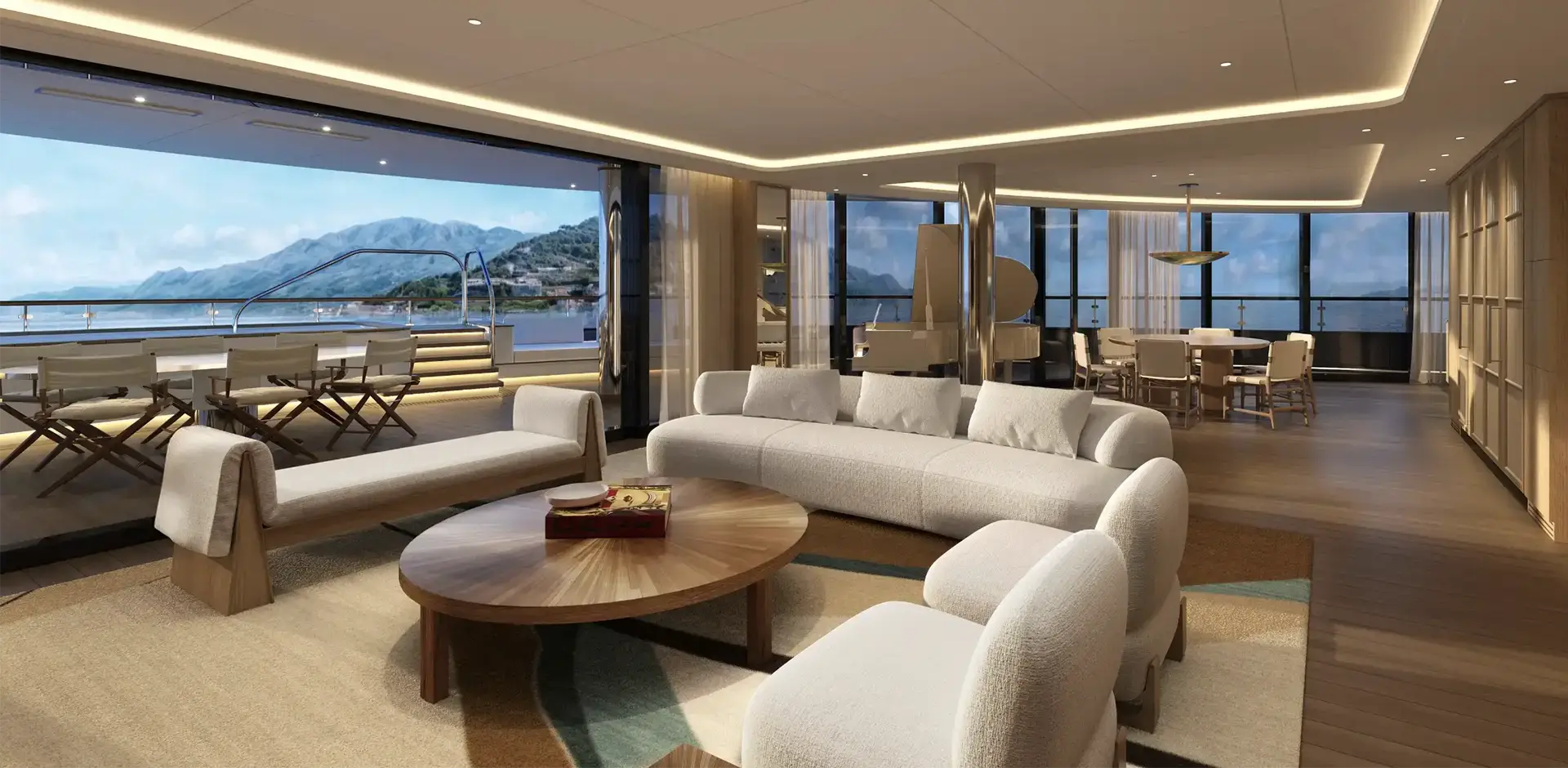 Luxurious open-plan lounge with white Sofas, a round wooden coffee table, and panoramic ocean and mountain views through floor-to-ceiling windows. Outside terrace features white chairs and railing.