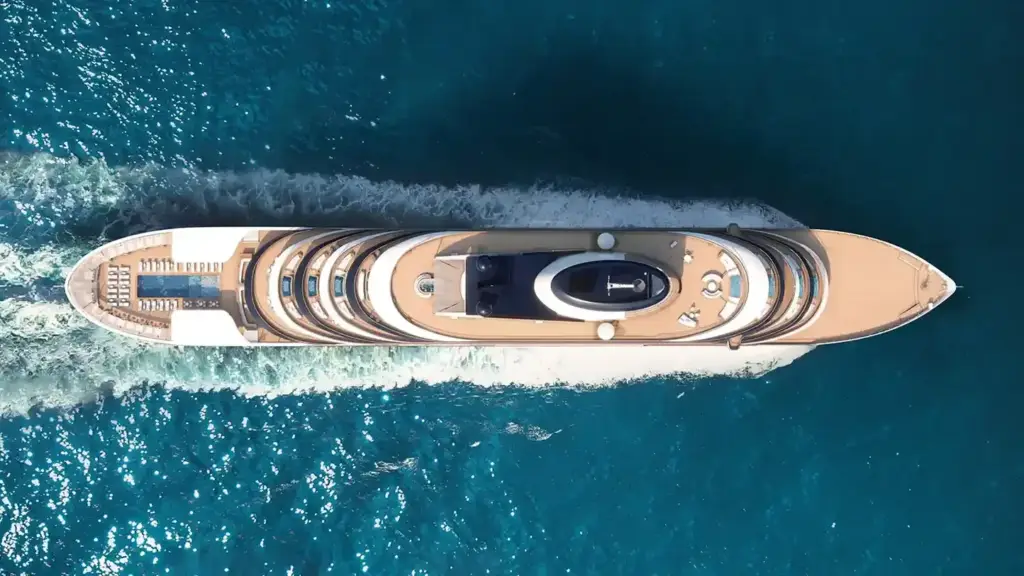 Aerial view of a large beige luxury yacht cruising through blue/teal ocean with a white wake behind it.