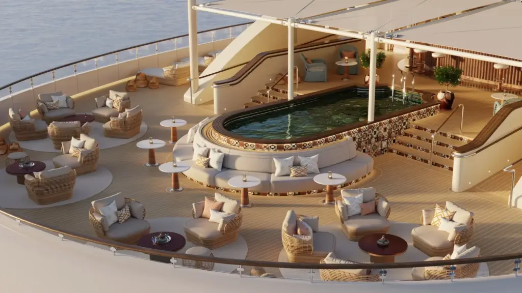 Open-air luxury cruise ship pool deck with a curved tiled pool, wicker lounges, and round white-topped tables arranged around cushioned seating; ocean visible on the left.