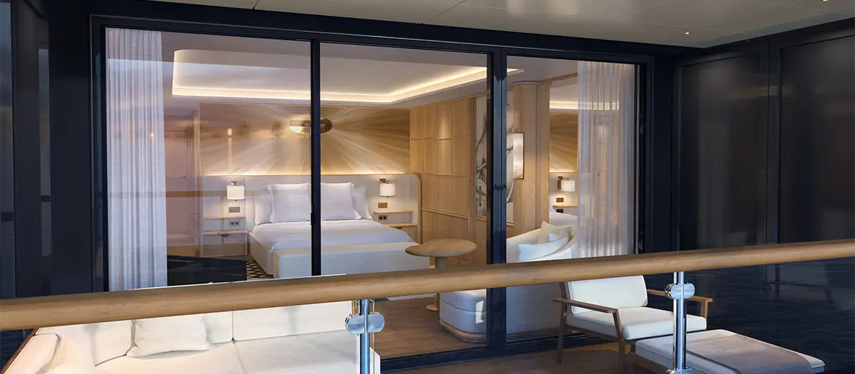Modern hotel suite seen through glass sliding doors, with a king bed, seating area, and soft ambient lighting on a private balcony.