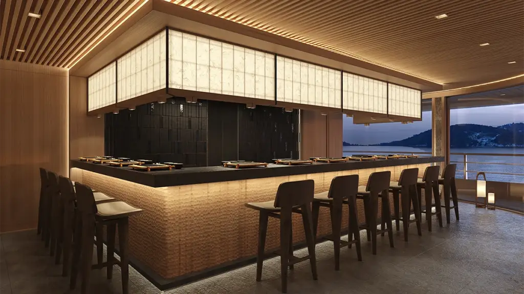 Elegant waterfront bar with a warm copper-toned counter, dark tile back wall, and row of wooden stools under soft hanging lights, overlooking the water.