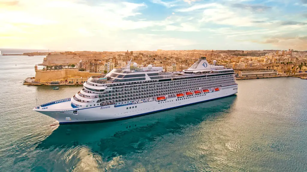 Large white cruise ship named Riviera docked in a harbor beside a historic walled city coastline at sunset.
