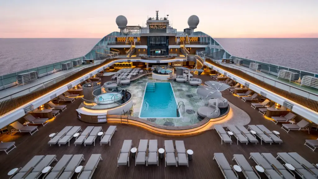 Sunset over a luxury cruise ship pool deck with rows of sun loungers and seating around a central pool.