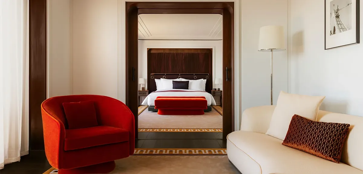 Living area of a luxury hotel suite: cream sofa with a brown pillow and a red curved chair, opening to a bedroom through dark wood arches.
