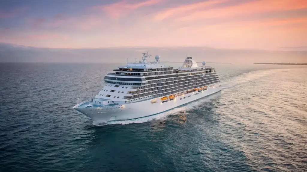 Large white cruise ship sailing on calm ocean at sunset with pink and orange sky nearby.