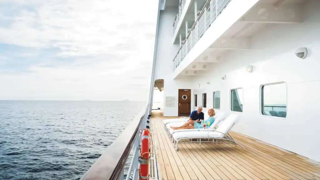 Couple lounging on white deck chairs on a cruise ship, sea to the left and bright sky above.