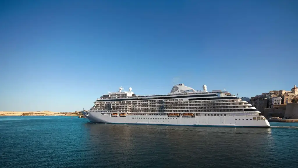 Large white cruise ship anchored in a calm harbor with a historic town along the shore under a clear blue sky.