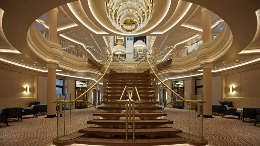 Luxurious hotel lobby with a grand curved wooden staircase and glass railings beneath a large crystal chandelier.