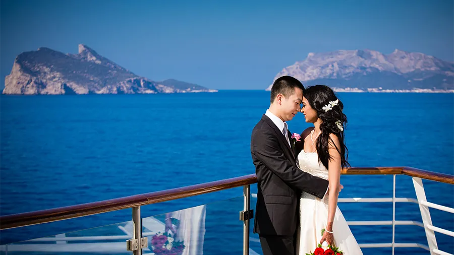 Bride in a White Wedding Dress and Groom in a Black Suit Embrace on a Deck by the Blue Ocean, Distant Mountains in the Background.