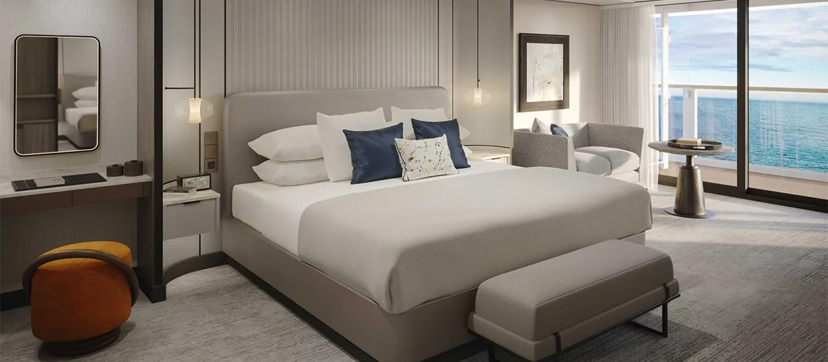 Hotel room with king bed, neutral decor, bench at foot, and ocean-view balcony beyond glass doors.