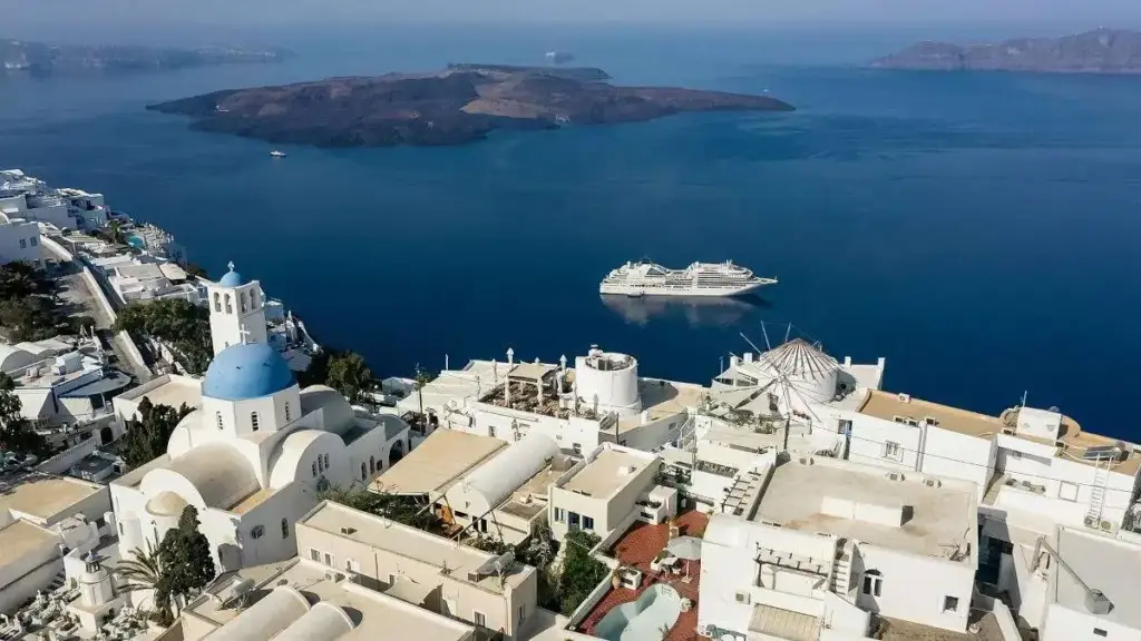 White Cycladic buildings and blue-domed church domes on a cliffside village overlooking the deep blue Aegean Sea; a cruise ship anchored nearby.
