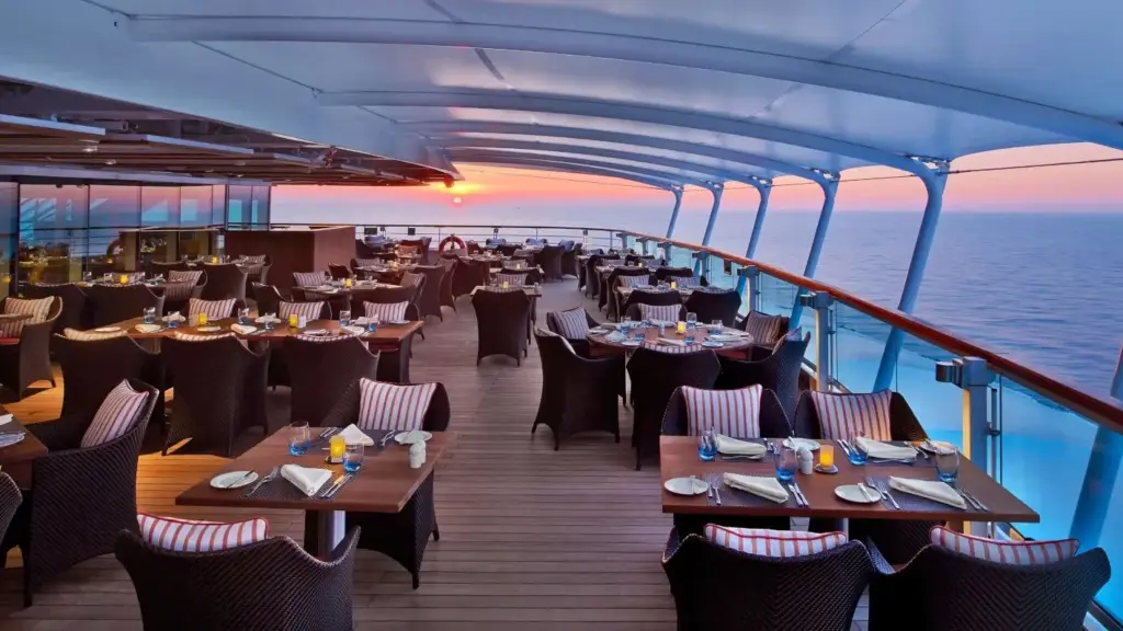 Open-air cruise ship restaurant deck at sunset with arranged tables and wicker chairs overlooking the sea.