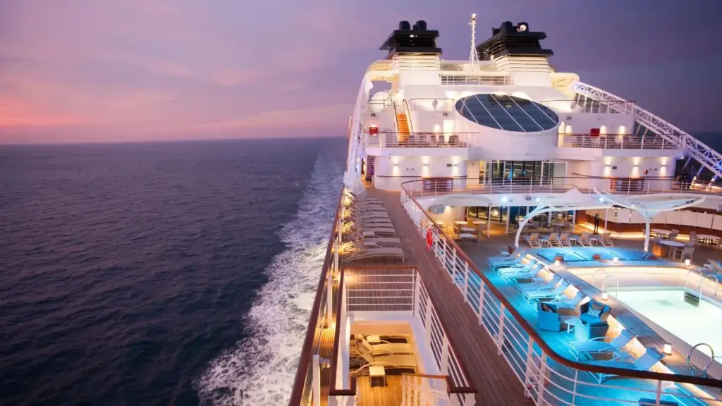 Sunset cruise ship deck with a row of lounge chairs, pool, and blue-lit pool area overlooking calm oceans.