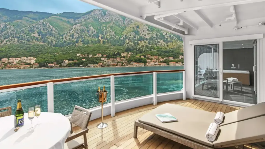 Balcony on a cruise ship with a lounge bed, glass sliding door to the cabin, and a table set for drinks overlooking blue water, hills, and a coastal town.