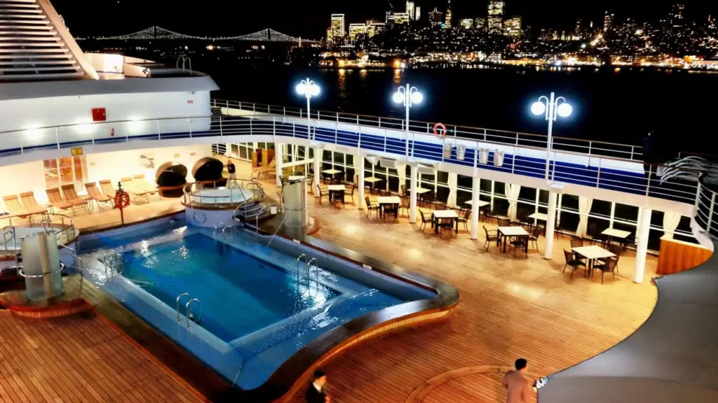 Night view of a cruise ship pool deck with wooden planks, lounge chairs, and tables; city skyline lights in the distance.