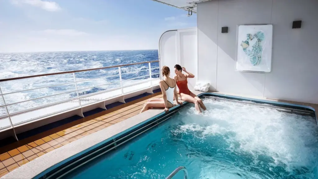 Two women in swimsuits sit on the pool edge of a cruise ship, chatting with the ocean rushing by nearby.